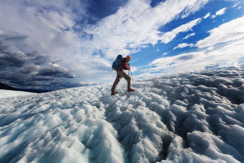 A person wearing climbing gear and a backpack is hiking up a snowy, icy slope under a dramatic, partly cloudy sky.