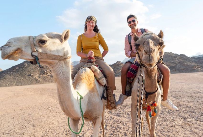 A happy honeymoon couple riding camels in a desert landscape with rocky hills in the background. Both are smiling and wearing casual clothes, with the woman on the left and the man on the right.