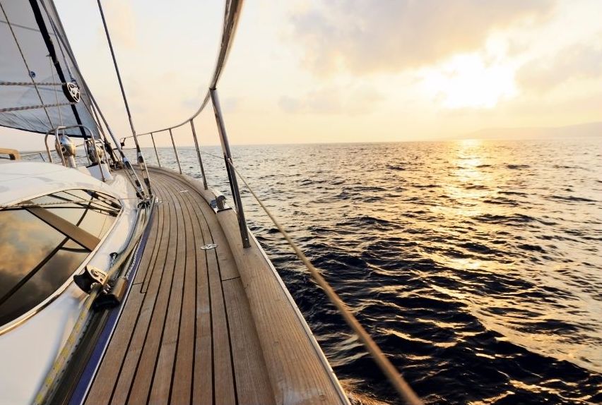 A sailboat glides on the ocean at sunset, with golden sunlight reflecting on the water and the boat’s wooden deck curving into the distance. The sky is partly cloudy, creating a peaceful and serene atmosphere.