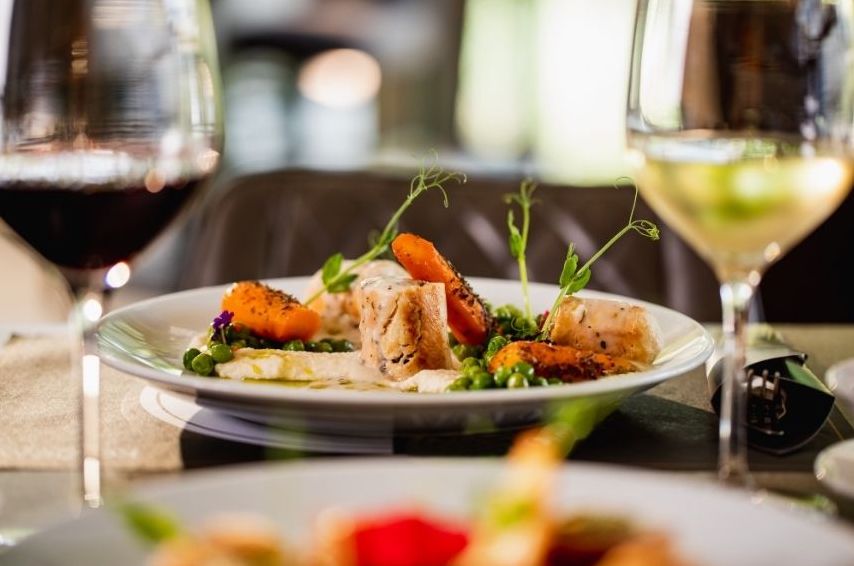 A close-up of an elegantly plated gourmet dish with vegetables and sauce, flanked by glasses of red and white wine on a dining table during one of our luxury golf vacations. The softly blurred background highlights the meal.