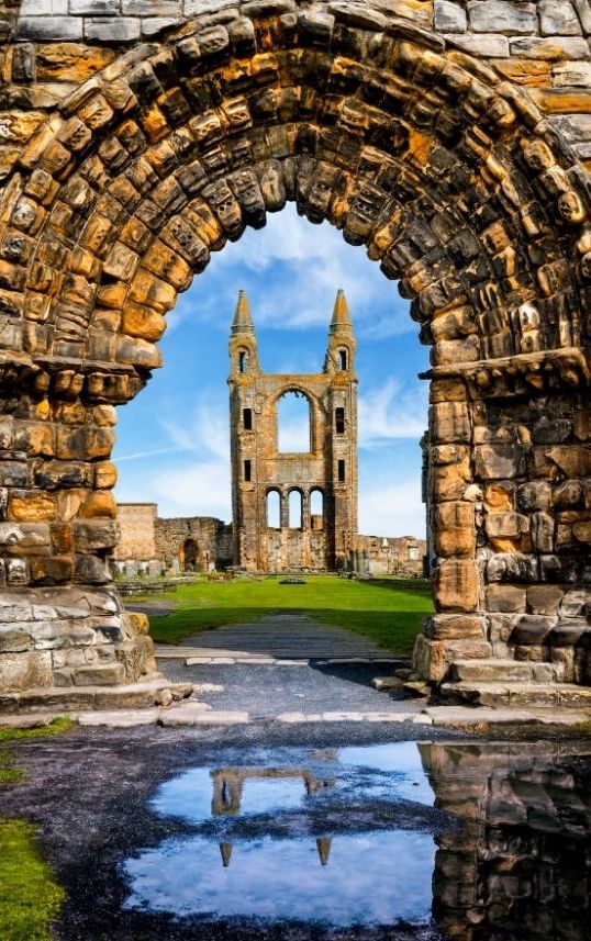 An arched stone doorway in St. Andrews Scotland frames the ruins of a gothic cathedral—an inspiring view for those enjoying luxury golf vacations. The historic site’s twin towers are reflected in a puddle, beneath blue sky and wispy clouds, surrounded by lush grass.
