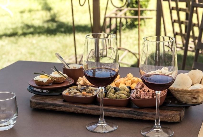 Two glasses of red wine sit on a table with a wooden board holding assorted cheeses, olives, bread rolls, spreads, and small appetizers—a perfect culinary spread for a gourmand, set outdoors with lush greenery in the background.