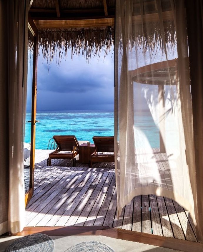 View from inside a bungalow through open curtains, showing two wooden lounge chairs on a deck overlooking clear blue ocean water under a cloudy sky.