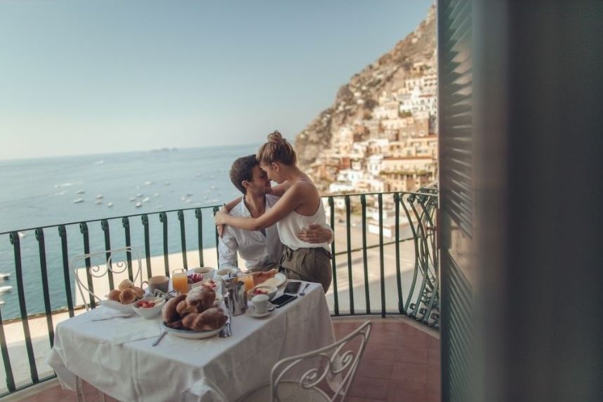 A happy honeymoon couple sits at a balcony table with breakfast, embracing and kissing, overlooking the sea and a coastal town built on a hillside with boats on the water below.