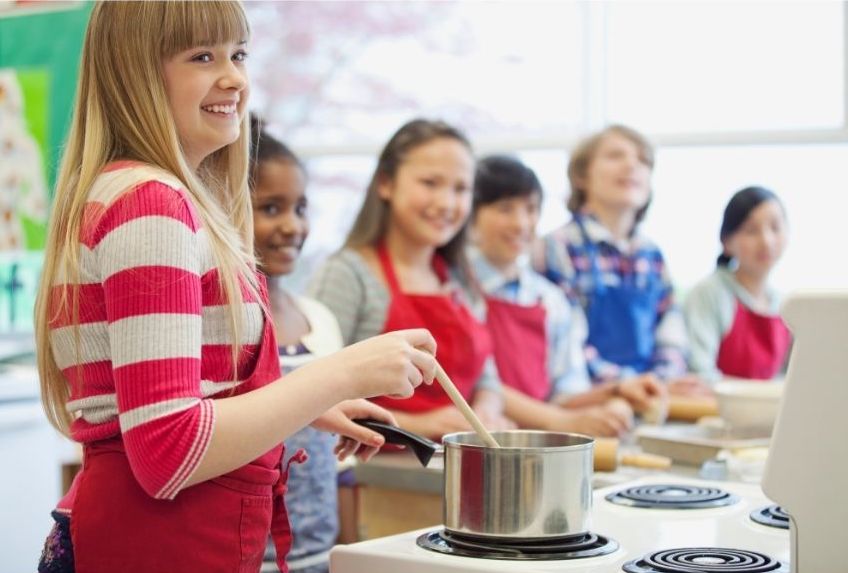 A group of smiling children wearing red aprons stand in a kitchen classroom. One girl in the foreground stirs a pot on the stove, while the others watch like eager culinary students in a fun cooking class.