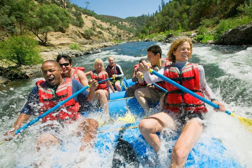 group of six people wearing red life jackets and holding paddles raft down a fast-moving river surrounded by trees and hills, smiling and splashed by water.