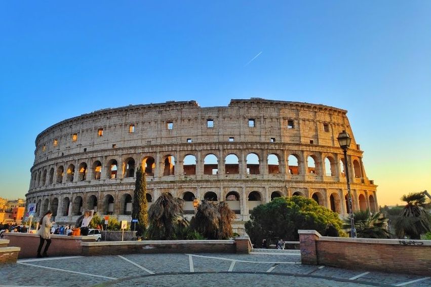 The Colosseum in Rome, Italy, is shown at sunset with a clear blue sky. People are walking and taking photos in the foreground, and trees are visible around the ancient amphitheater.