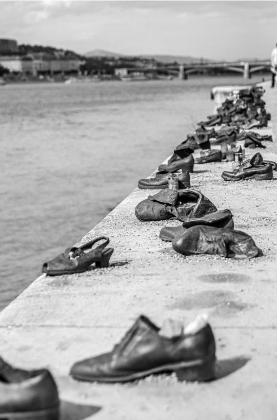 Rows of old, empty shoes are lined up along the edge of a stone embankment by a river, a poignant WWII memorial with the city and bridge in view—an essential stop for history travel and reflection.