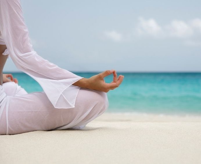 A woman in white clothing sits cross-legged on sand near the sea, doing yoga.