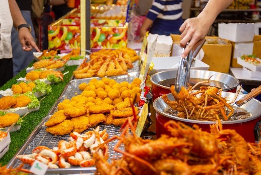 A variety of fried seafood and breaded snacks displayed at a market stall; a culinary enthusiast uses tongs to pick up fried prawns from a pan, with other golden-brown food items arranged on trays nearby.