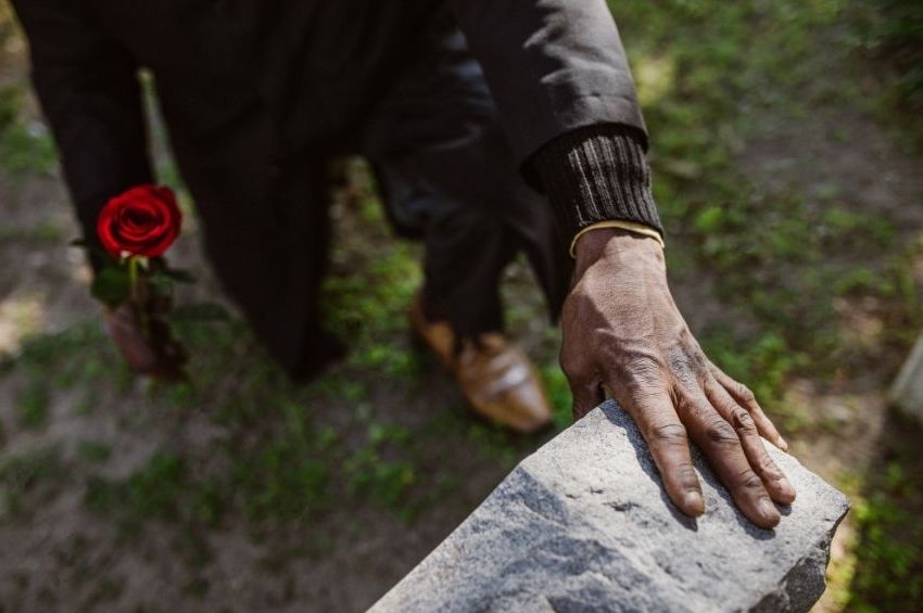 A person in dark clothing holds a red rose in one hand while touching a gravestone with the other, standing on grass at an outdoor historical site—a poignant moment during a WWII history travel tour.