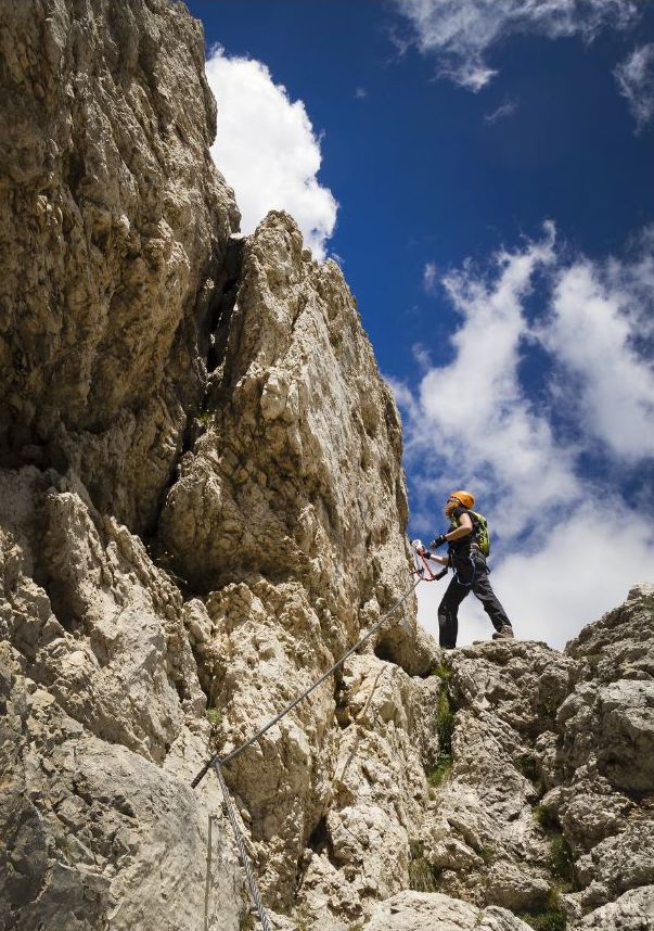A climber wearing a helmet and carrying a backpack ascends a rocky mountain, using a secured climbing rope against a backdrop of blue sky and white clouds.