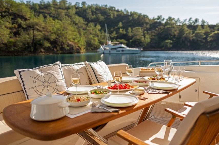 A dining table on a private yacht deck is set with plates, salads, wine glasses, and food, overlooking calm water and a forested shoreline in Croatia with another yacht in the distance under a clear sky.