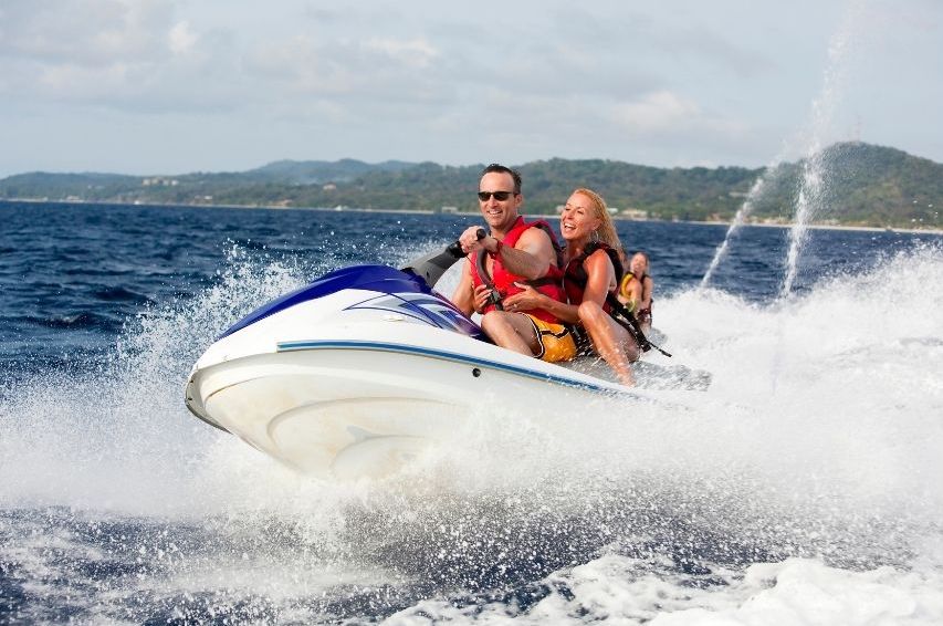 A man and a woman ride a jet ski together, smiling and enjoying themselves on the water near Croatia’s scenic coastline, while another person follows behind. The ocean waves splash around, with private yachts visible in the distance.