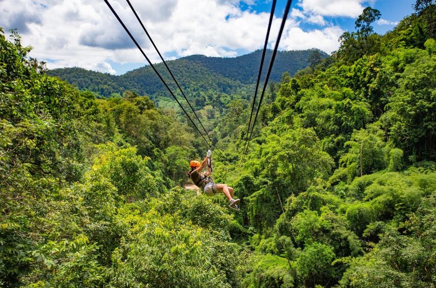 A person in a harness ziplining high above a lush, green forest with rolling hills and a partly cloudy sky in the background.